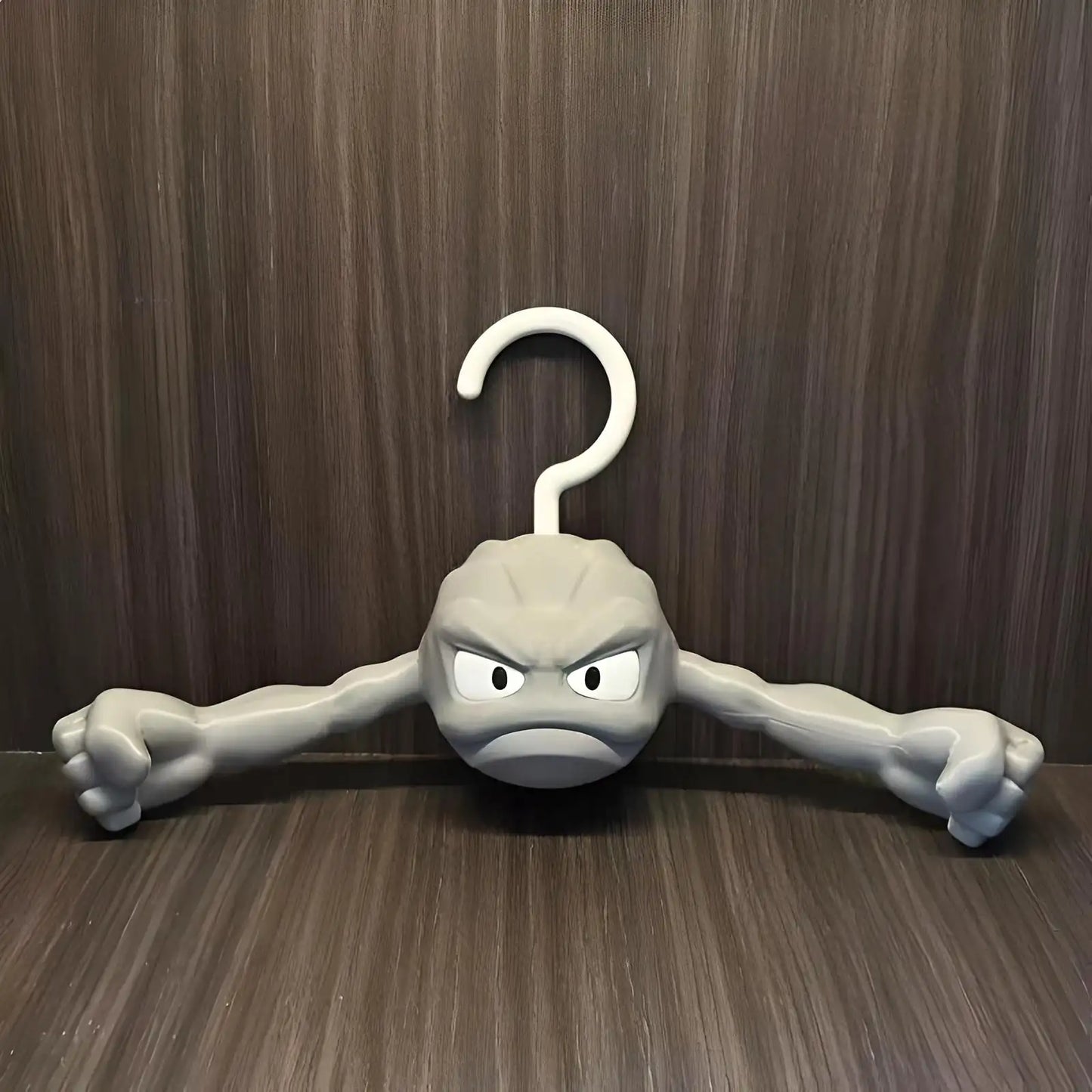 Anime-inspired Geodude clothes hanger with flexing rock arms against a dark wood wall background.