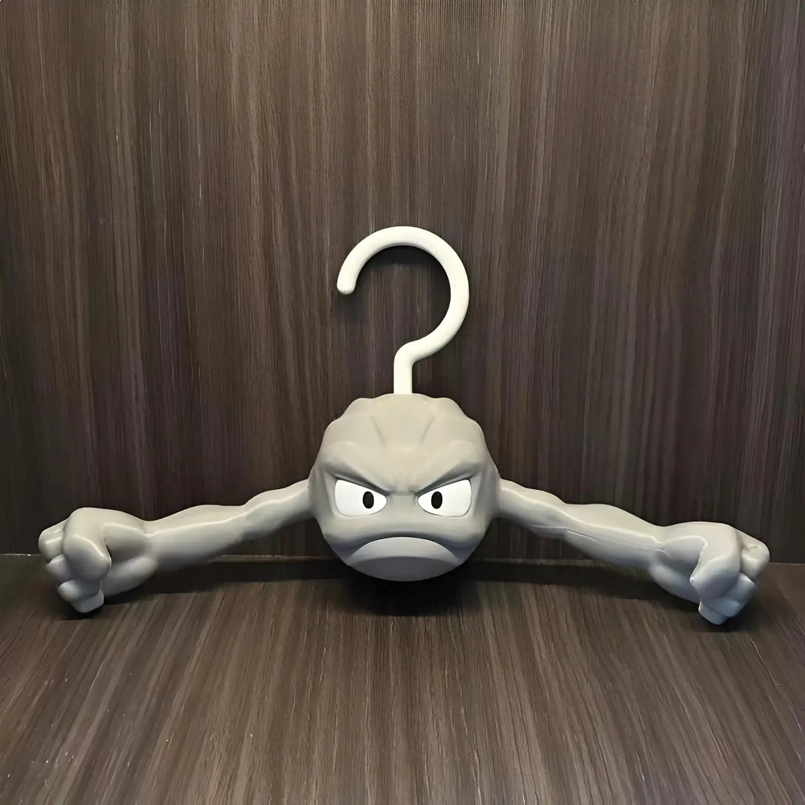 Anime-inspired Geodude clothes hanger with flexing rock arms against a dark wood wall background.